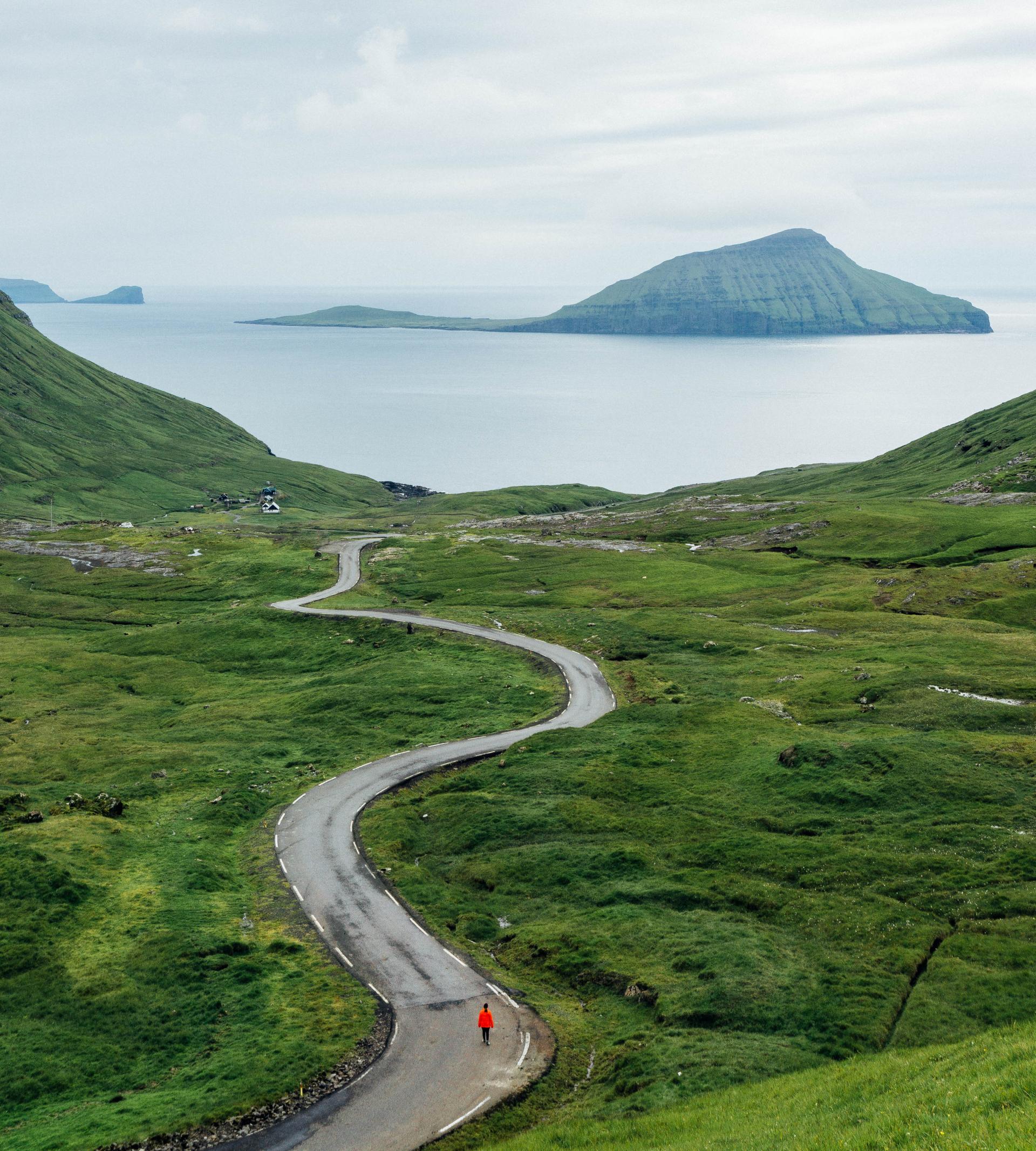 Thumbnail of - Man in red jacket, walking on a scenic road in Nordadalur, Faroe Islands. 